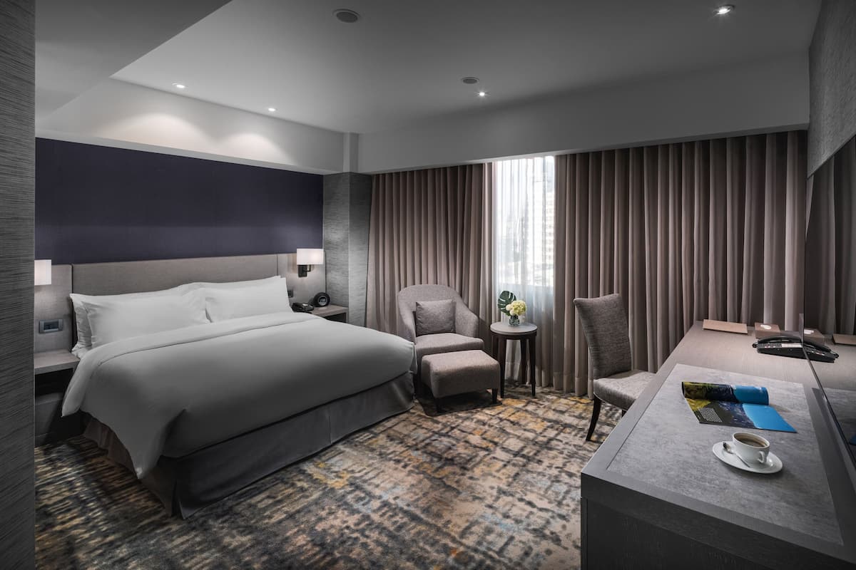 Une chambre d’hôtel moderne avec un grand lit, un bureau avec un ordinateur portable, une chaise et une petite table sur laquelle se trouvent une tasse et des fleurs.