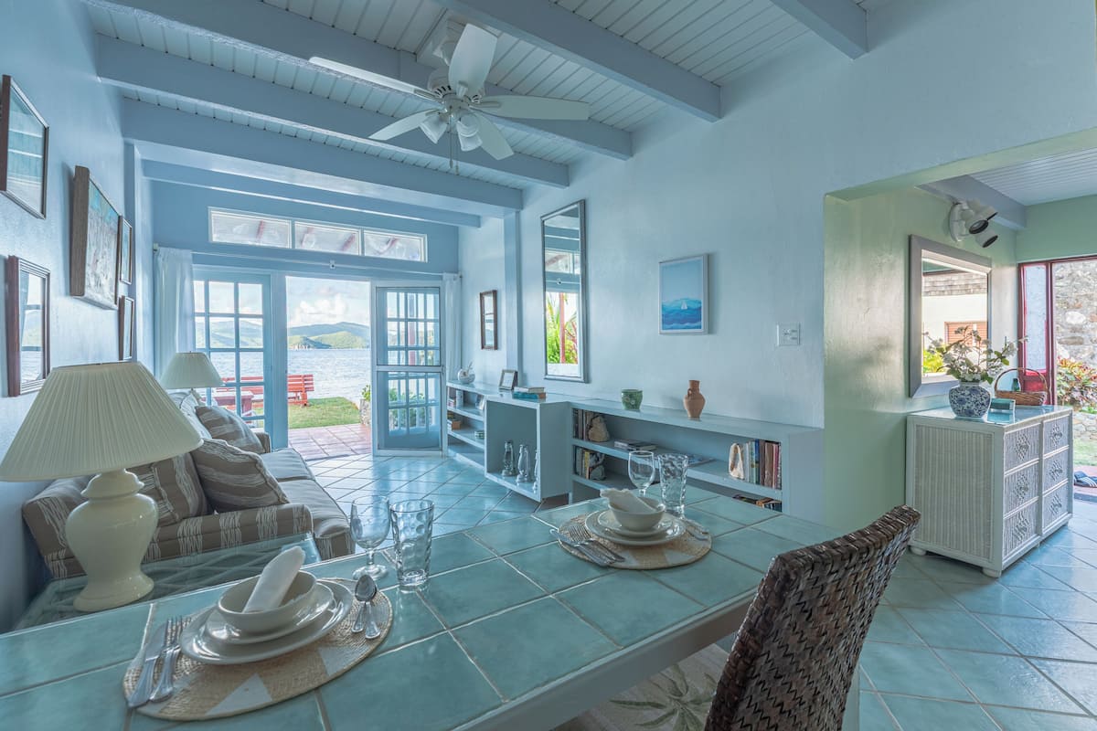 A spacious dining area with a view of the sea, a ceiling fan, and a dining table set with plates and glasses.