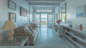 A living room with a sofa, a glass coffee table, a lamp, a bookshelf, and a view through a glass door. - Fort Recovery Beachfront Resort (West End)