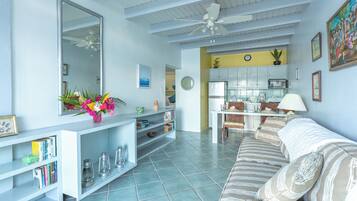 A living room with a sofa, a coffee table, a bookshelf, a dining area, and a kitchen.