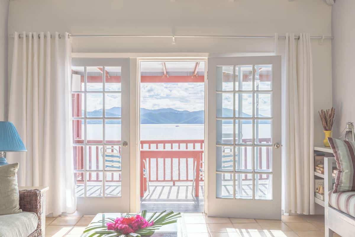 A room with a view of mountains through a large window, a balcony with a red railing, and a wicker sofa.