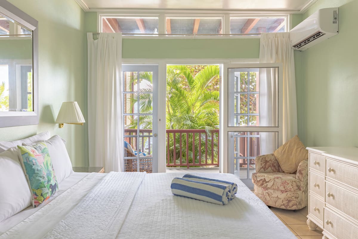A bedroom with a bed, a chair, a dresser, and a balcony with a view of palm trees.