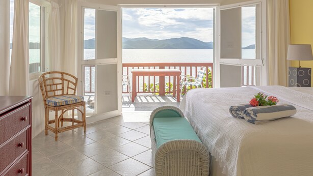 A bedroom with a bed, a chair, a dresser, and a view of a balcony with a railing. - Fort Recovery Beachfront Resort (West End)