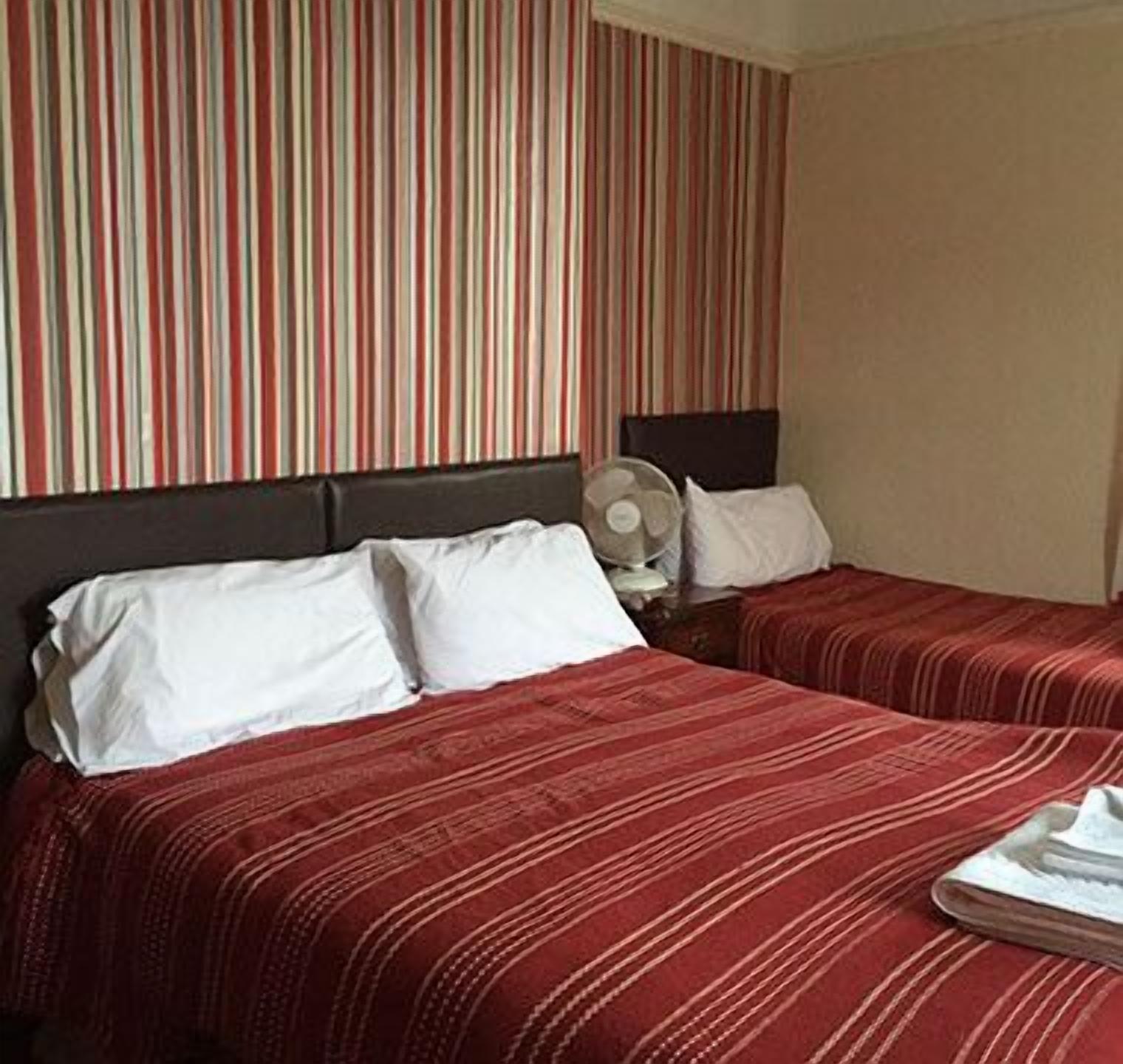 A hotel room with two beds, striped curtains, a bedside table with a fan, and a white pillow.