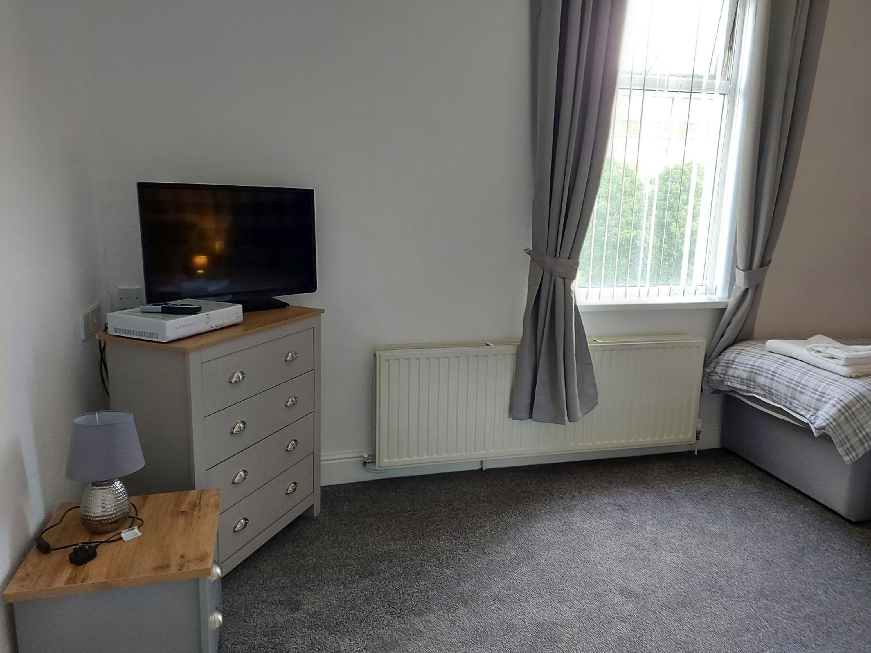 Ein Schlafzimmer mit Fernseher auf einer Kommode, einem Heizkörper, einem Fenster mit Vorhängen und einem Bett mit Decke.