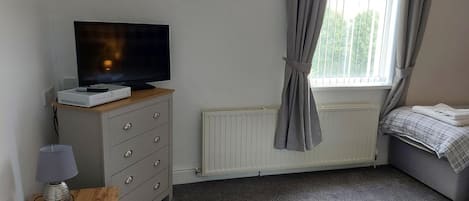 Ein Schlafzimmer mit Fernseher auf einer Kommode, einem Heizkörper, einem Fenster mit Vorhängen und einem Bett mit Decke.