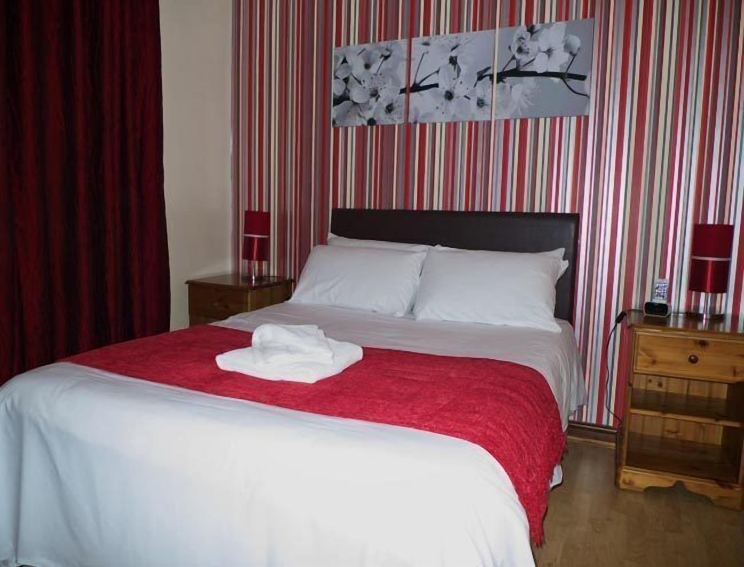 Un lit bien fait, avec du linge de lit blanc et une couverture rouge, deux tables de chevet avec des lampes, et un rideau à rayures en arrière-plan.