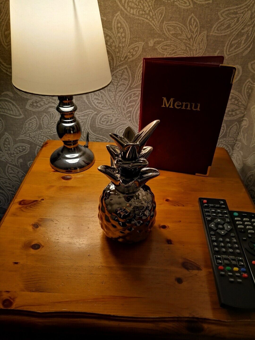 A wooden nightstand with a lamp, a pineapple-shaped salt and pepper shaker, a remote control, and a menu book.