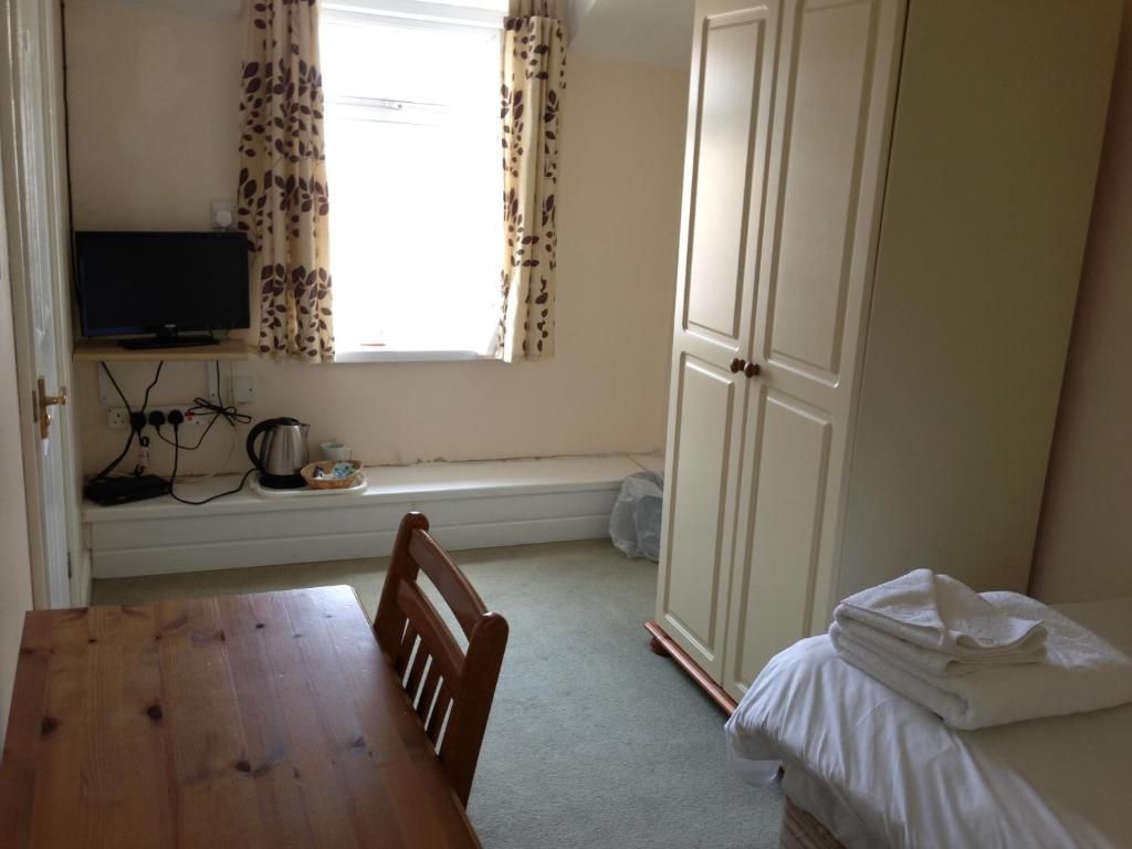 A room with a wooden table, a television on a shelf, a window with curtains, a wardrobe, and a bed with a folded white towel.