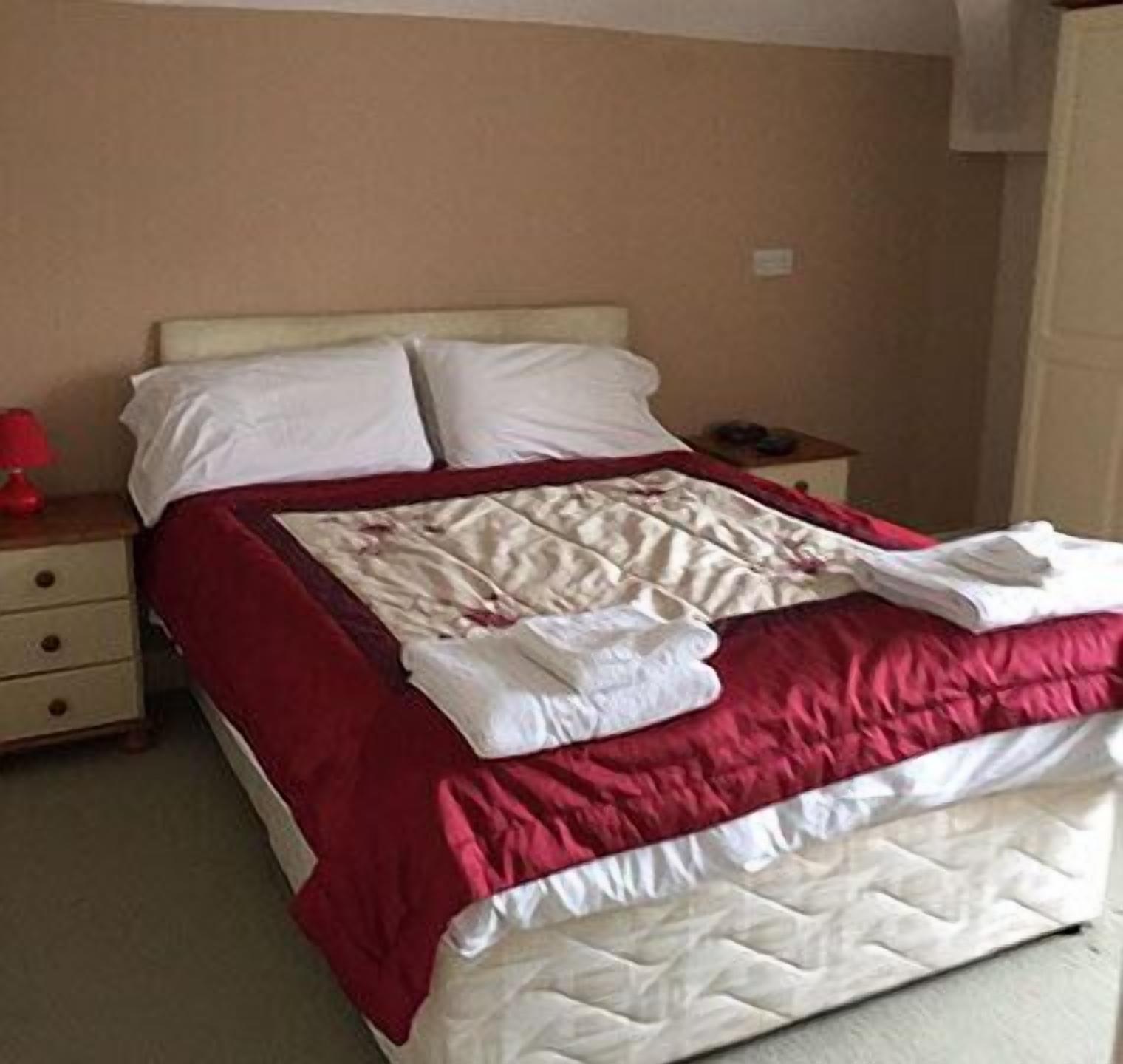 A neatly made bed with white and red bedding, a bedside table with a lamp, and a small nightstand with a phone.