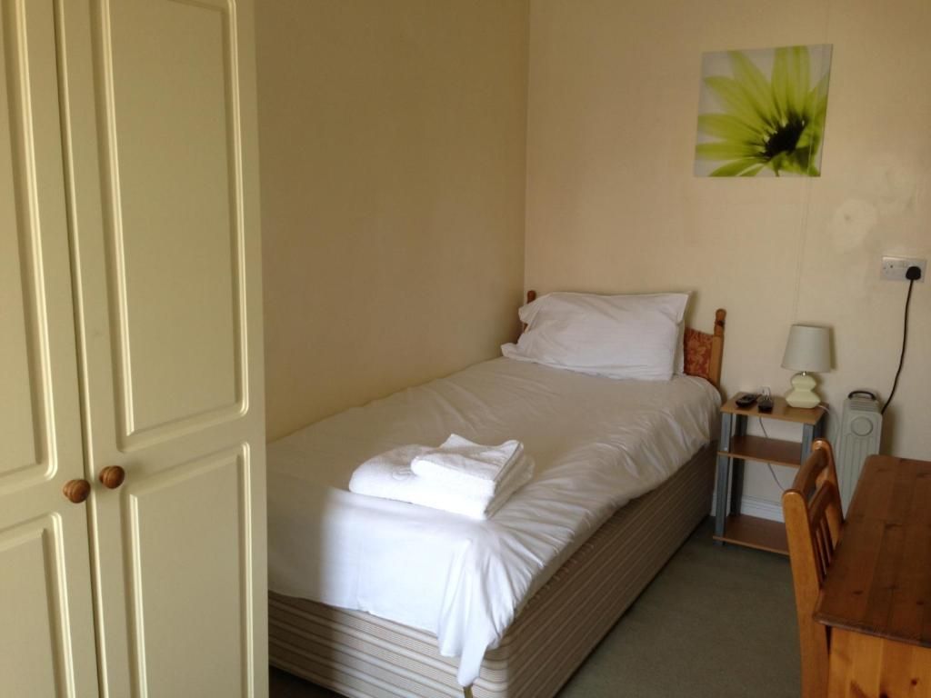 A neatly made bed with white linens and a folded towel, a bedside table with a lamp, and a wall-mounted power outlet.