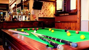 A pool table with balls and cues, a bar area with bottles and a TV, and wooden paneling on the walls.