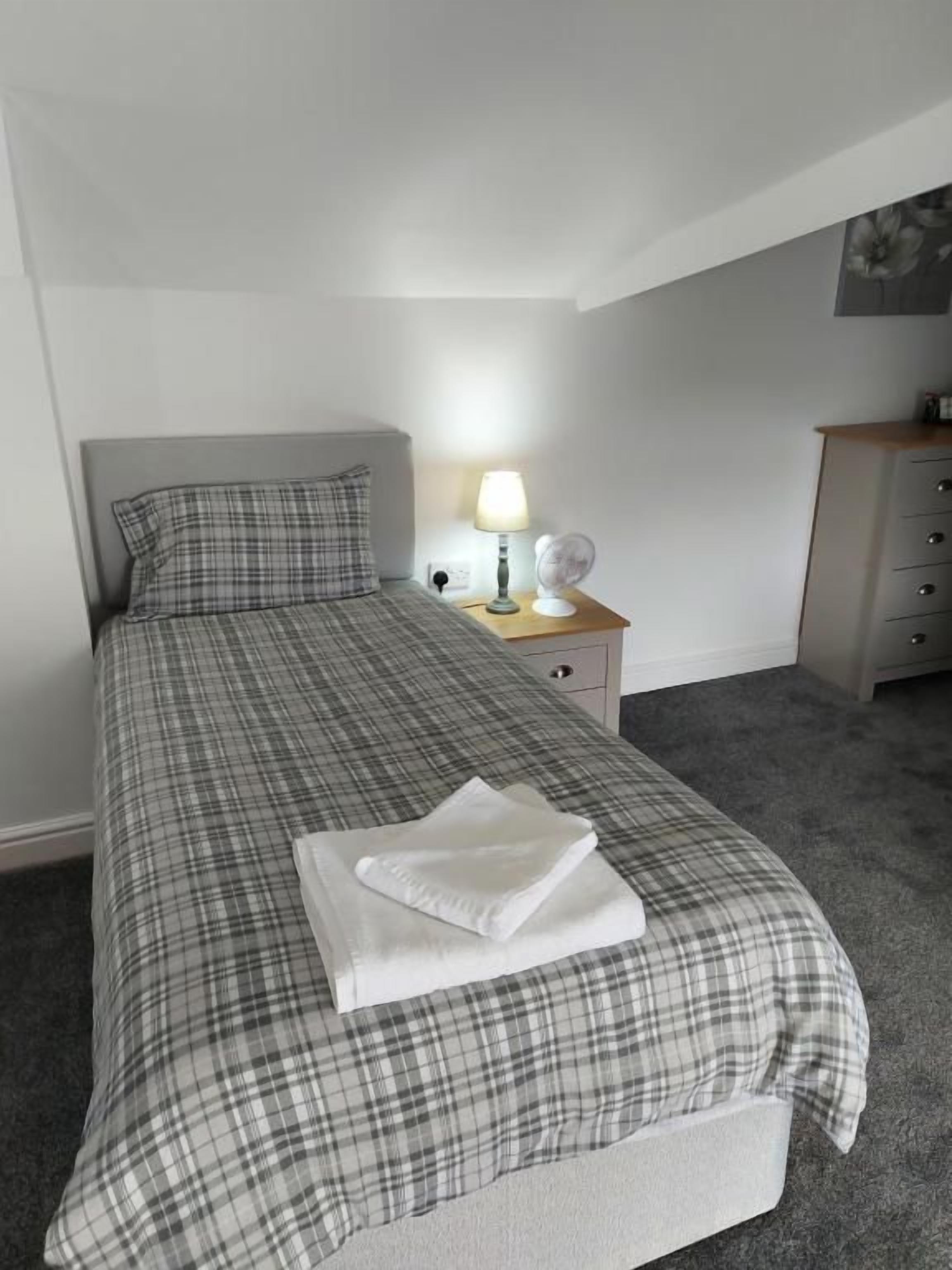 A neatly made bed with a plaid bedspread, a bedside lamp, and a small bedside table with a lamp and a fan.