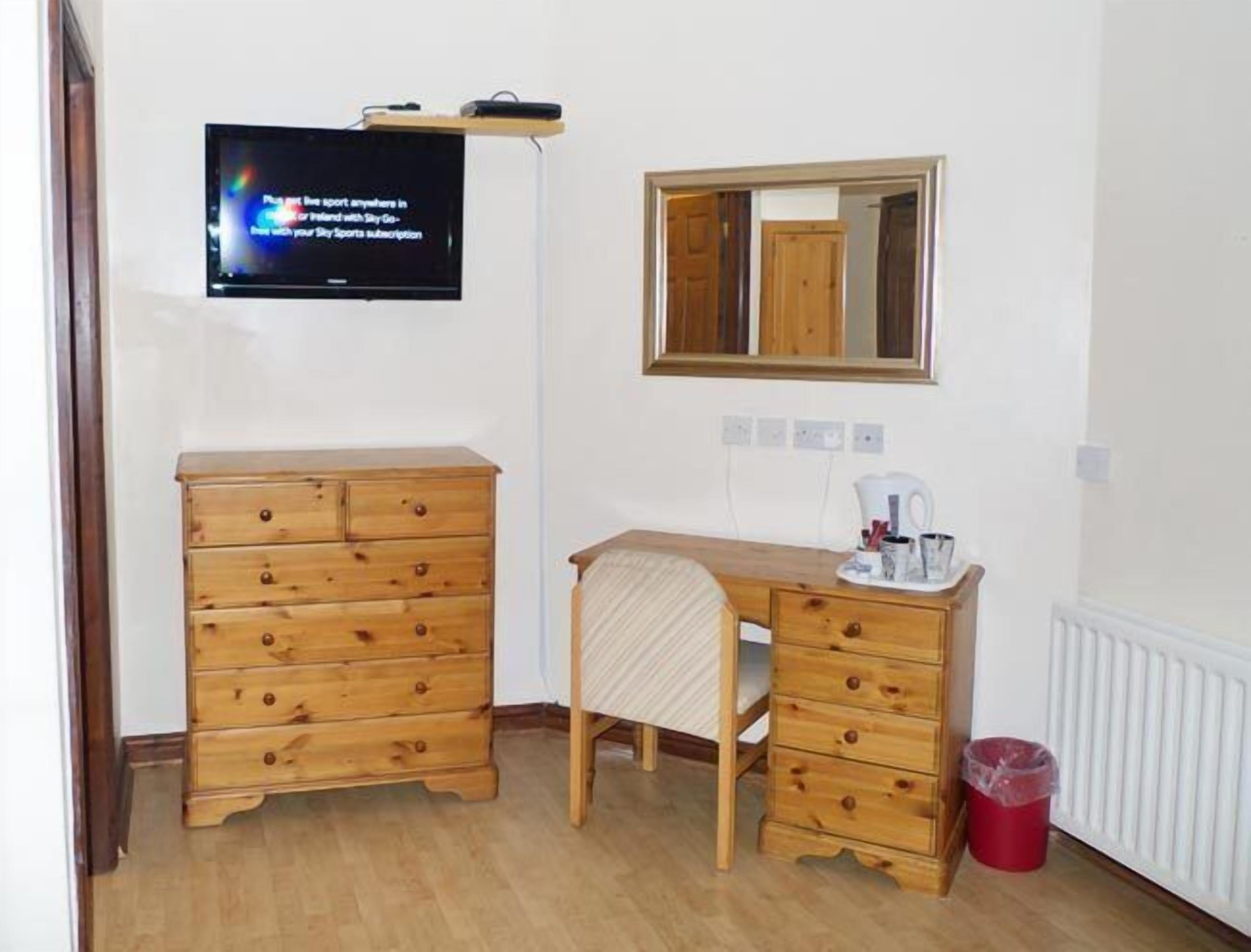 A room with a wooden dresser, a desk with a chair, a television mounted on the wall, and a mirror above the desk.