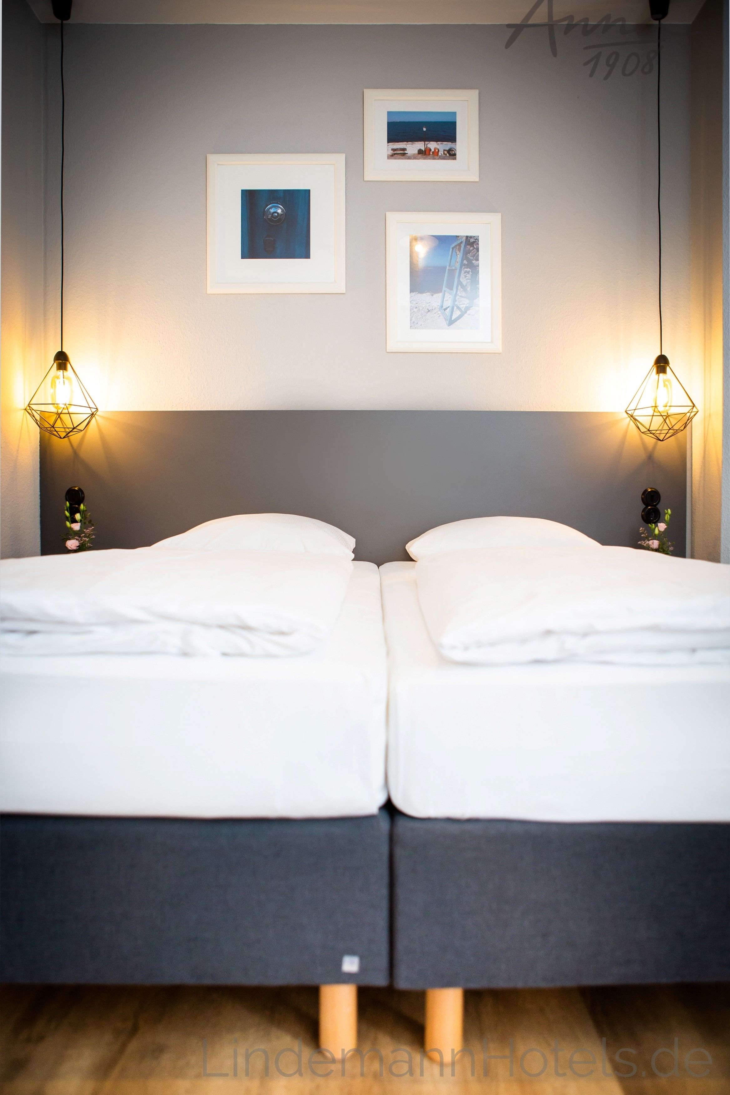 Ein Doppelbett mit grauem Kopfteil, zwei Wandlampen und drei gerahmte Bilder an der Wand.