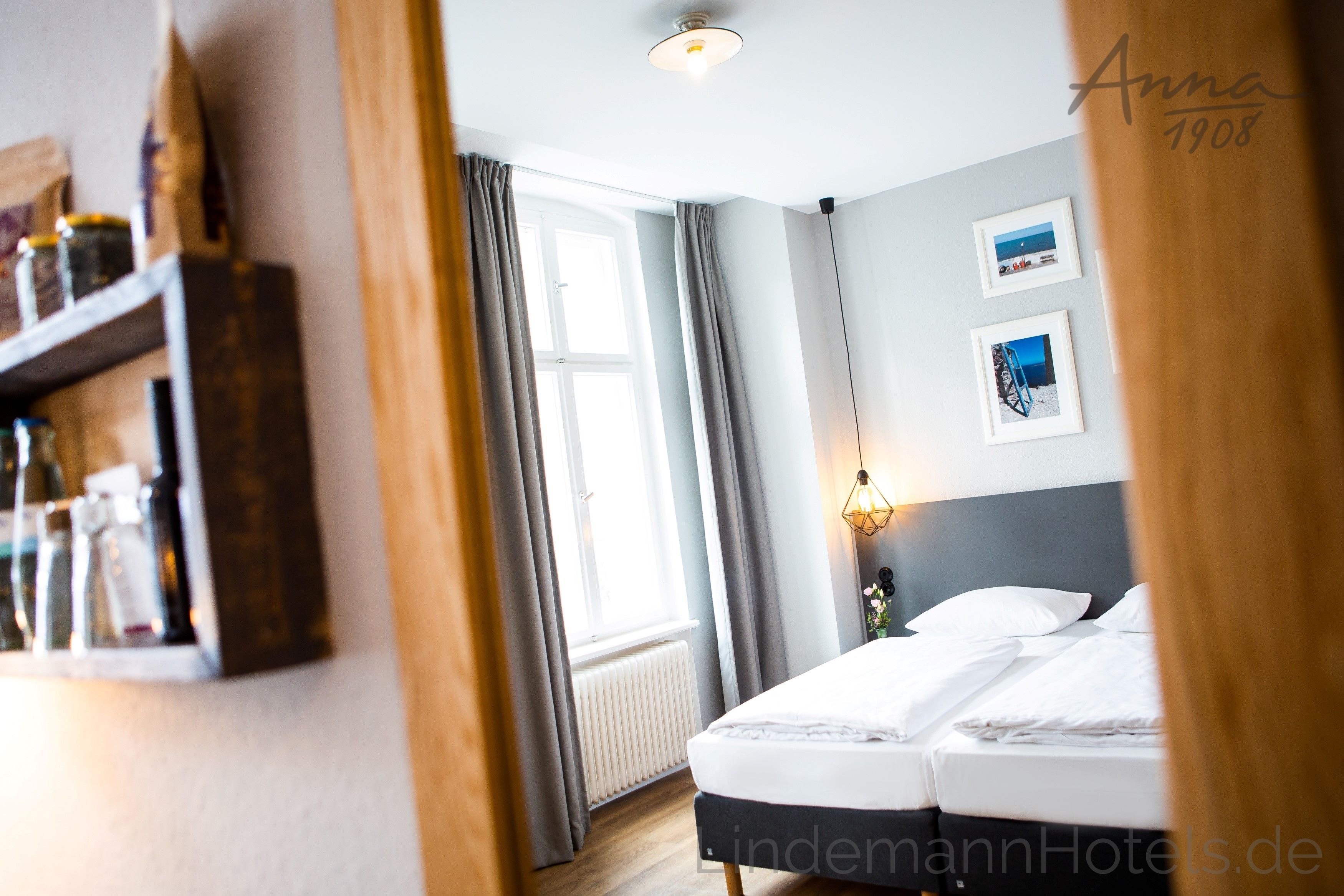 A hotel room with a bed, a window with curtains, a shelf with bottles, and framed pictures on the wall.