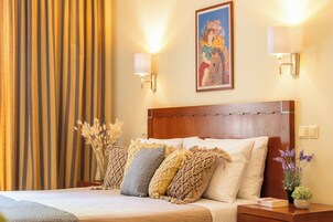 Habitación de hotel con cabecera de madera, cama con varios almohadones, mesita de noche con lámpara, cuadro enmarcado en la pared y cortina a rayas verticales.
