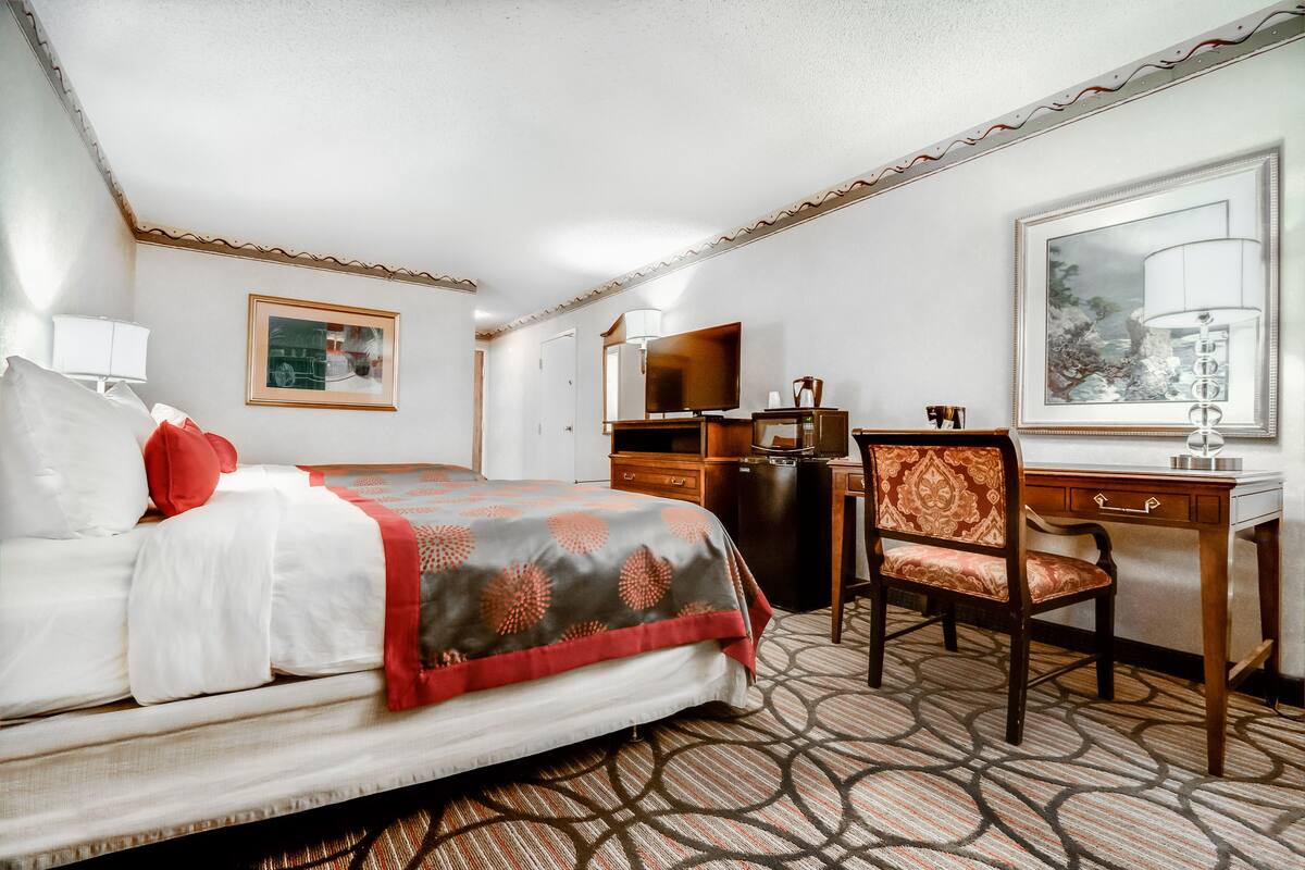 A hotel room with a bed, a desk, a chair, a dresser, a mirror, and a large window.