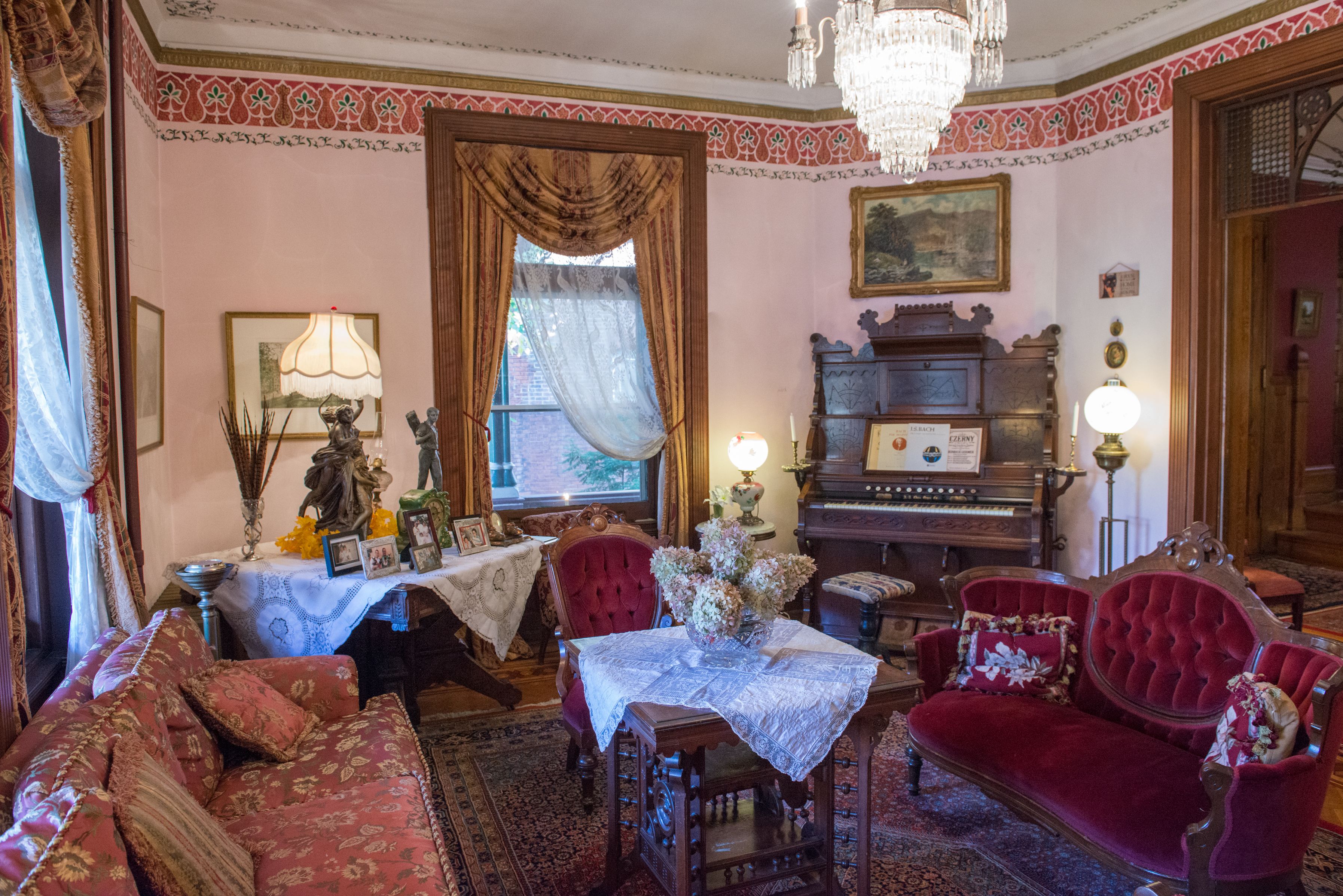 A vintage room with a red sofa, a wooden table with a lace tablecloth, a red armchair, a chandelier, and a piano.