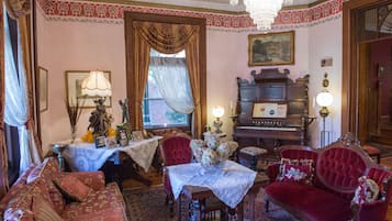 A vintage room with a red sofa, a wooden table with a lace tablecloth, a red armchair, a chandelier, and a piano.