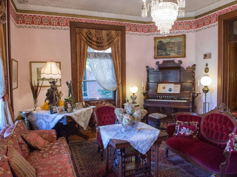 A vintage room with a red sofa, a wooden table with a lace tablecloth, a red armchair, a chandelier, and a piano.