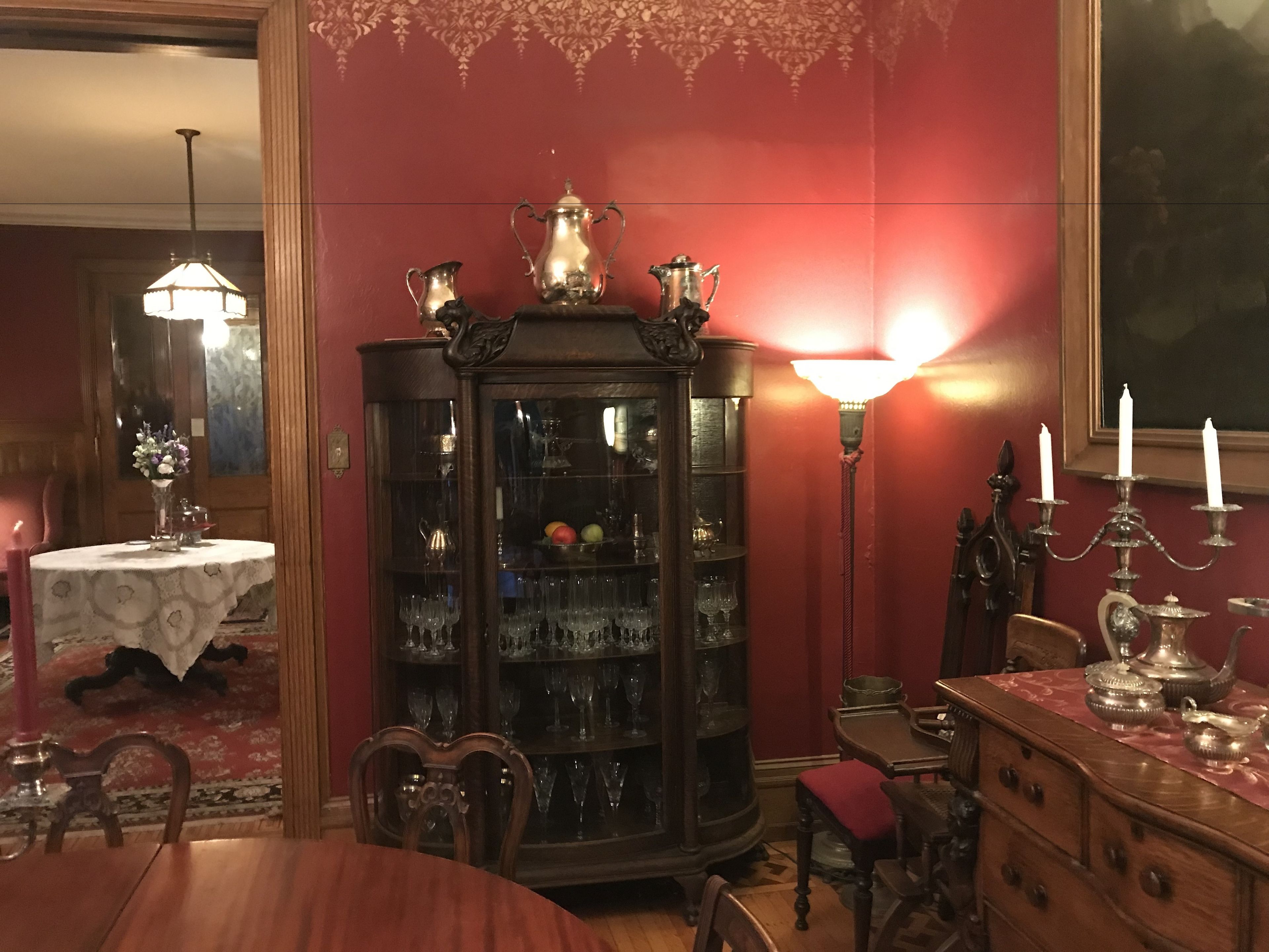 A dining room with a wooden table, chairs, a china cabinet with glassware, and a red wall.