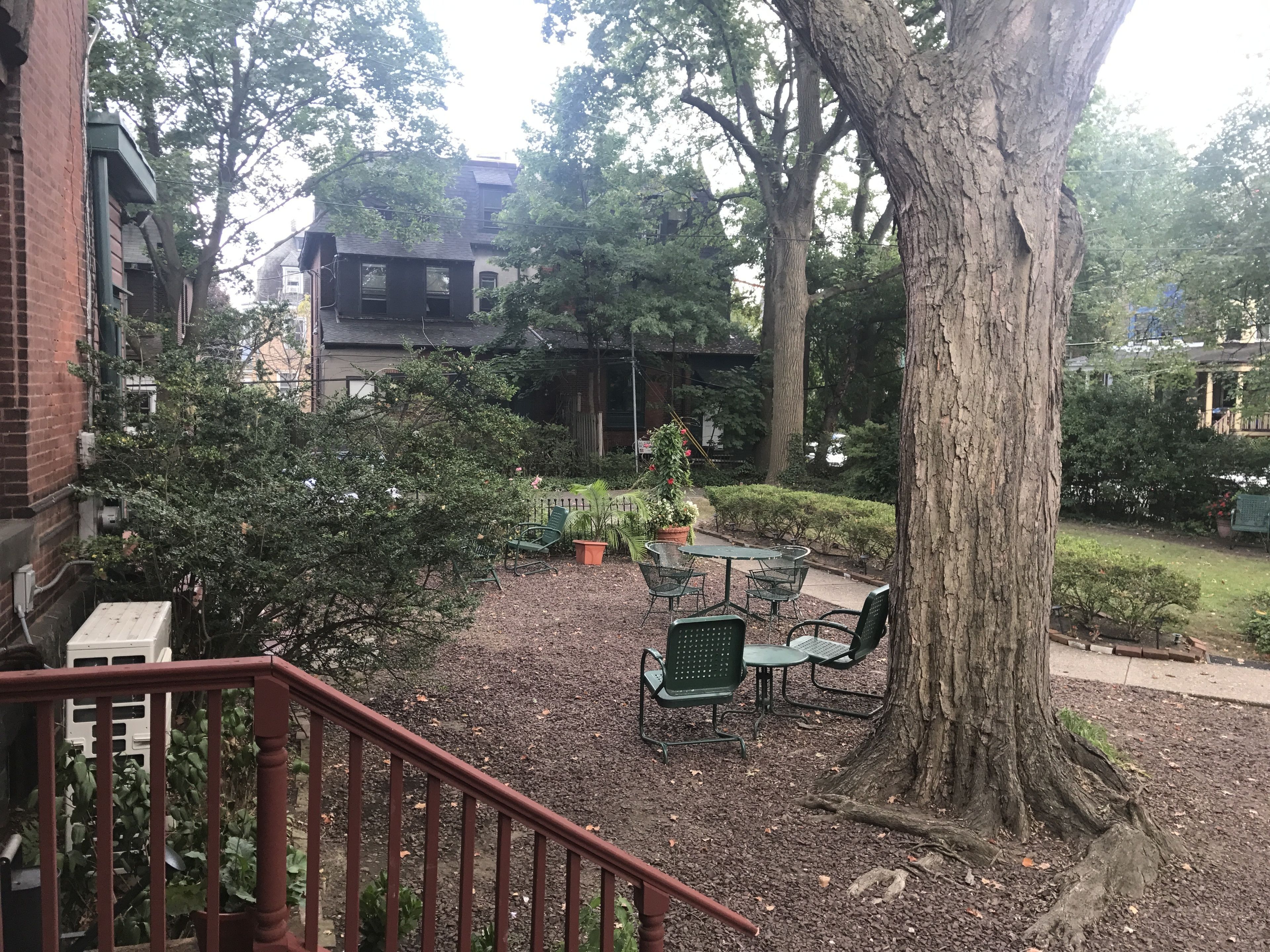 A courtyard with a wooden staircase, a table with chairs, and a large tree.
