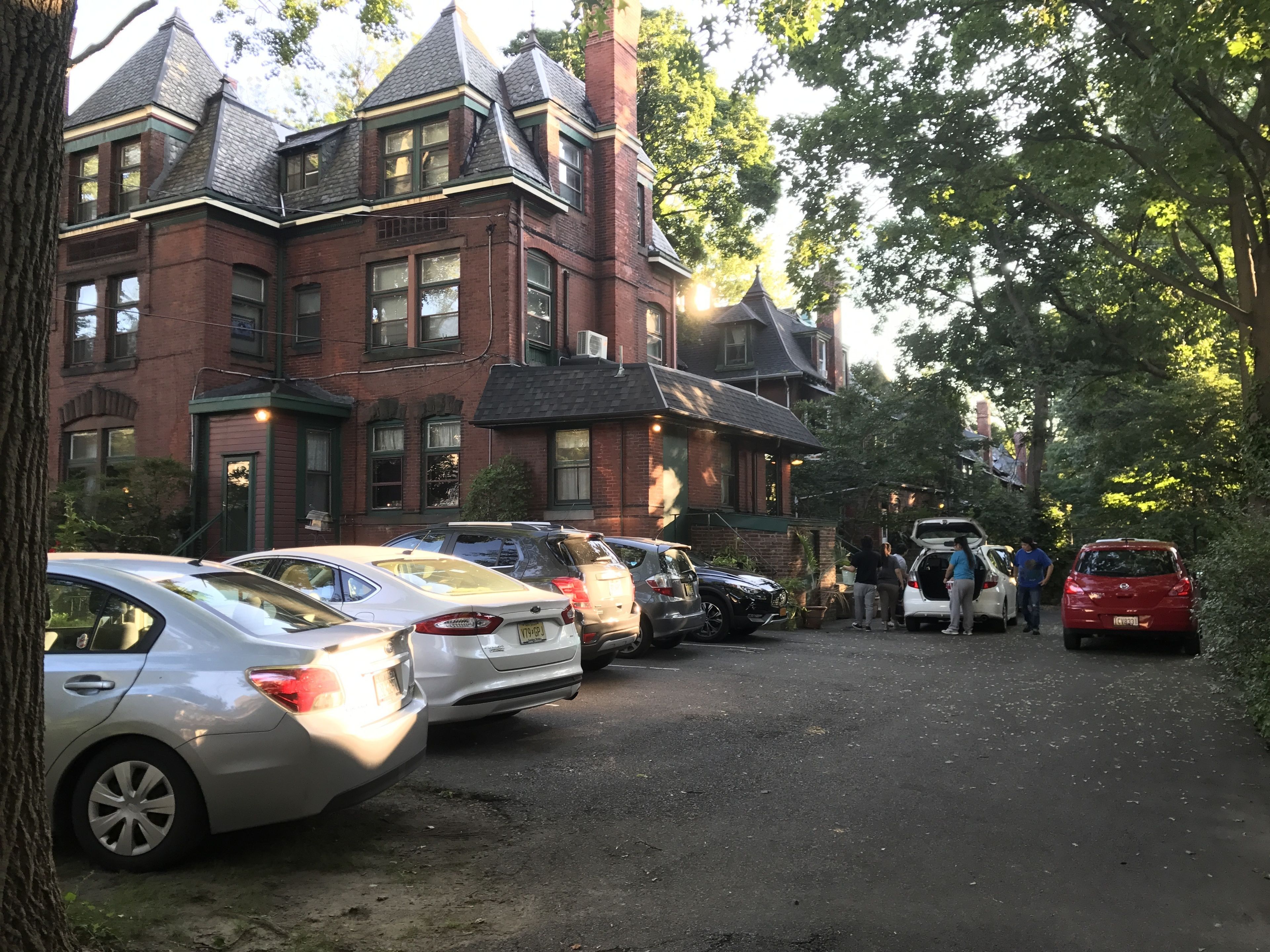 A row of parked cars in front of a large brick building with a steep roof and multiple windows.