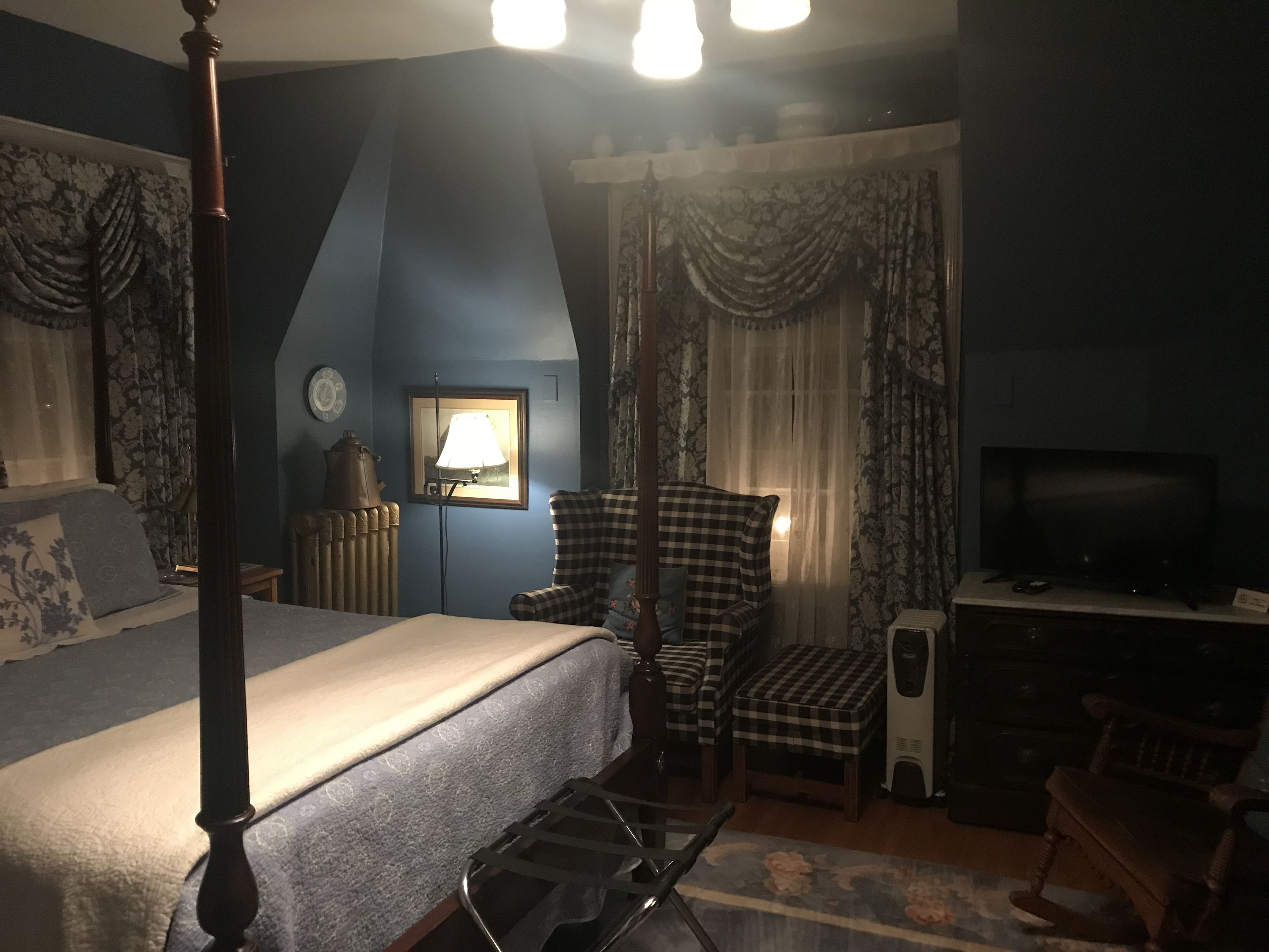 A hotel room with a four-poster bed, a checkered armchair, a television, and a radiator.
