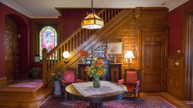 Un magnifique escalier orné d’un lustre, une table ronde agrémentée d’un vase de fleurs et un sol recouvert d’une moquette rouge.