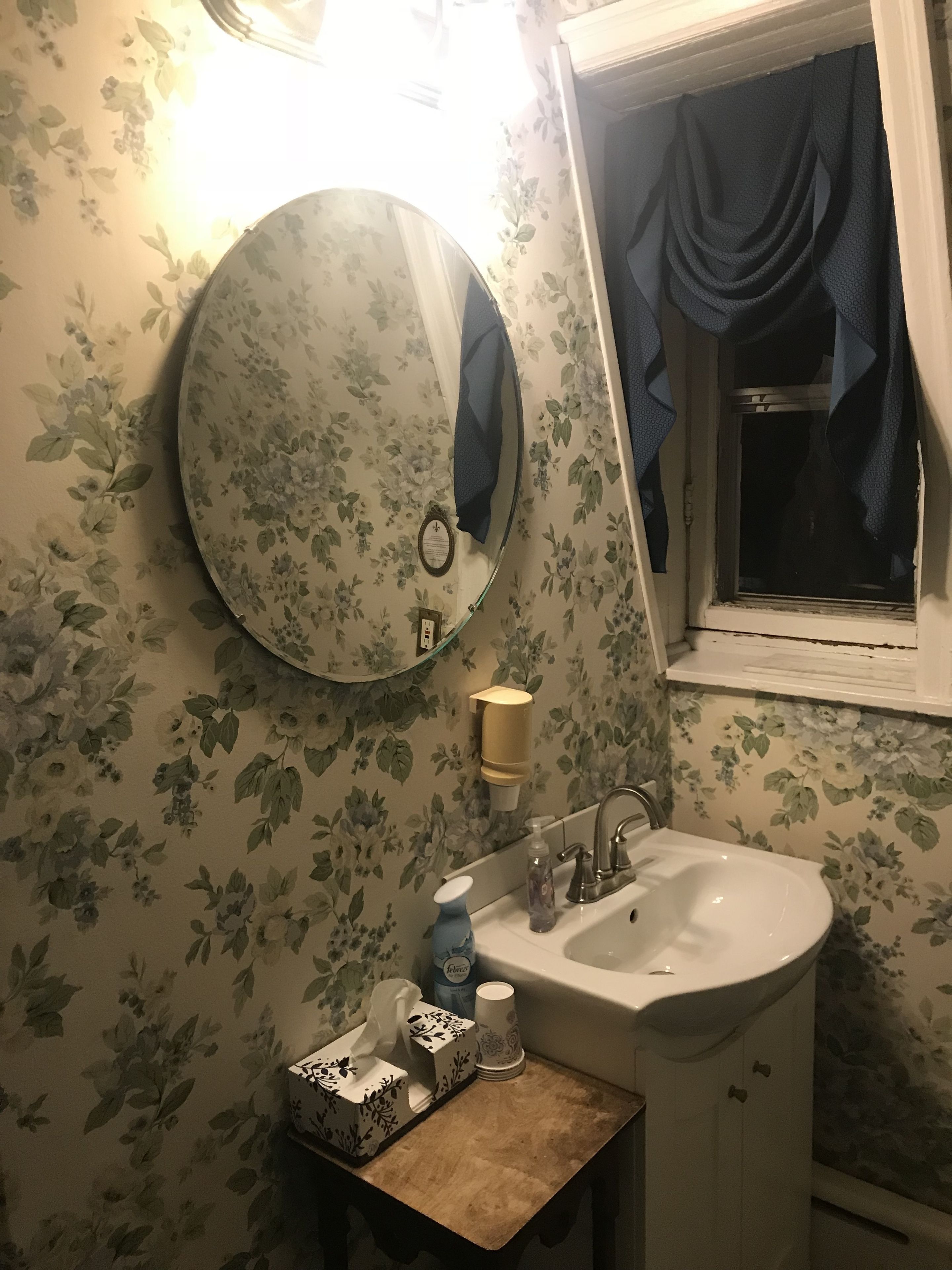 Uma casa de banho com papel de parede floral, um espelho redondo, uma pia, uma caixa de lenços e uma janela com cortina.