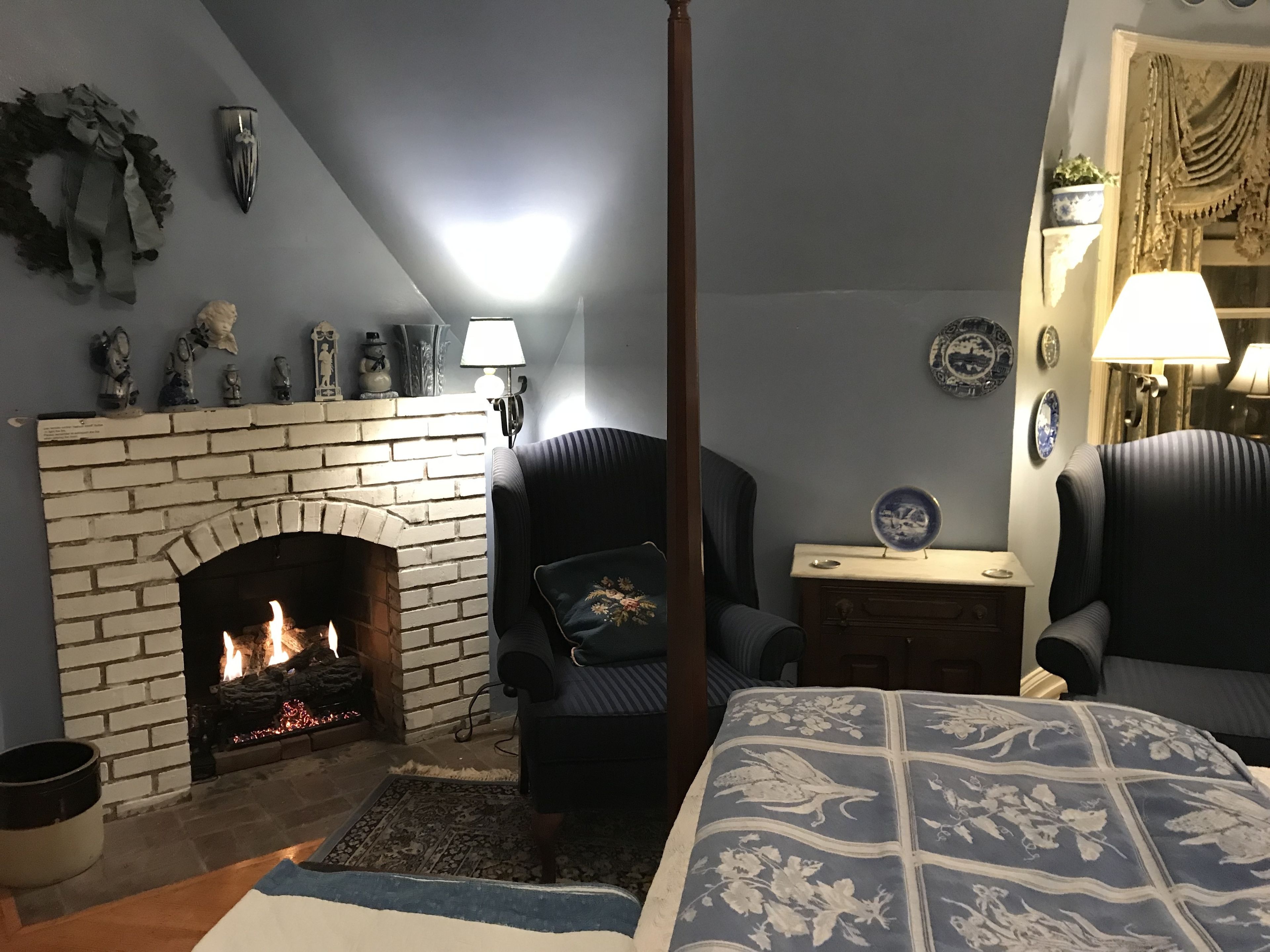 A cozy room with a brick fireplace, a comfortable armchair, a bedside table with a lamp, and a bed with a floral-patterned bedspread.