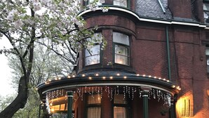 Une maison de style victorien dotée d’une baie vitrée en arc de cercle, de colonnes vertes et d’un porche décoré de guirlandes lumineuses.