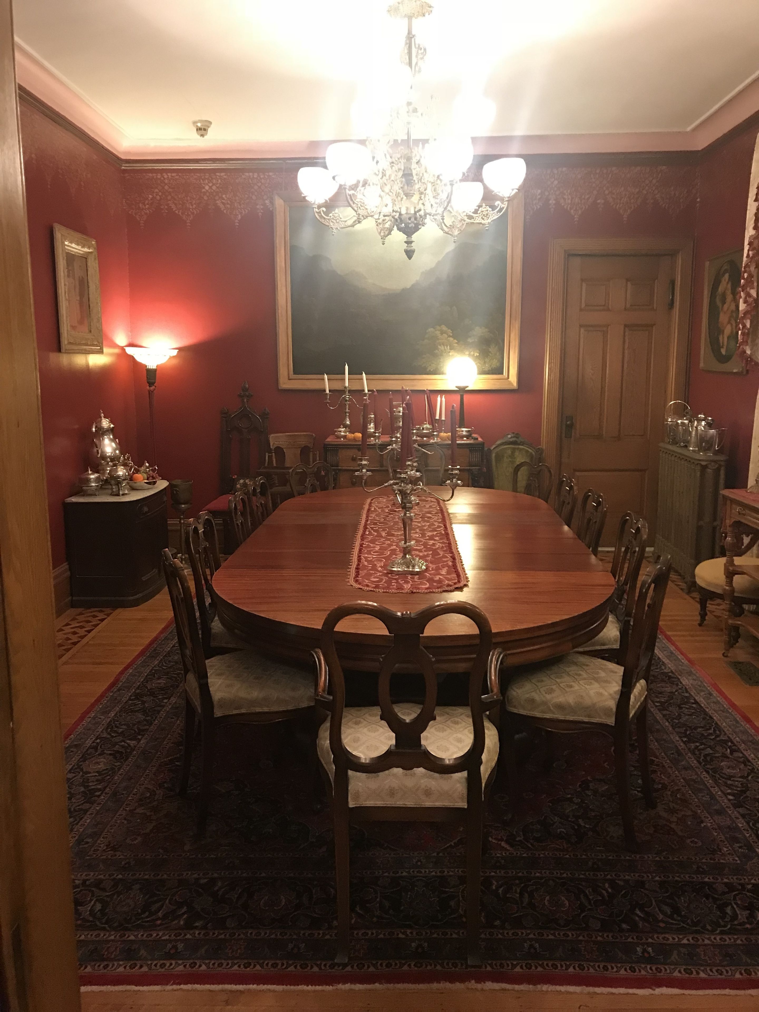 Uma sala de jantar formal com uma grande mesa de madeira, cadeiras ornamentadas, um candelabro e uma pintura de paisagem na parede.