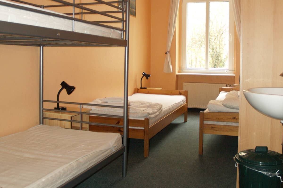 A dormitory room with bunk beds, a desk, a chair, a window with curtains, a radiator, and a sink.