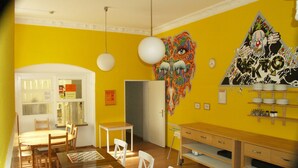 A dining area with a yellow wall, white chairs, a wooden table, and a wall mural. - HappyGoLucky Hotel & Hostel (Berlin)