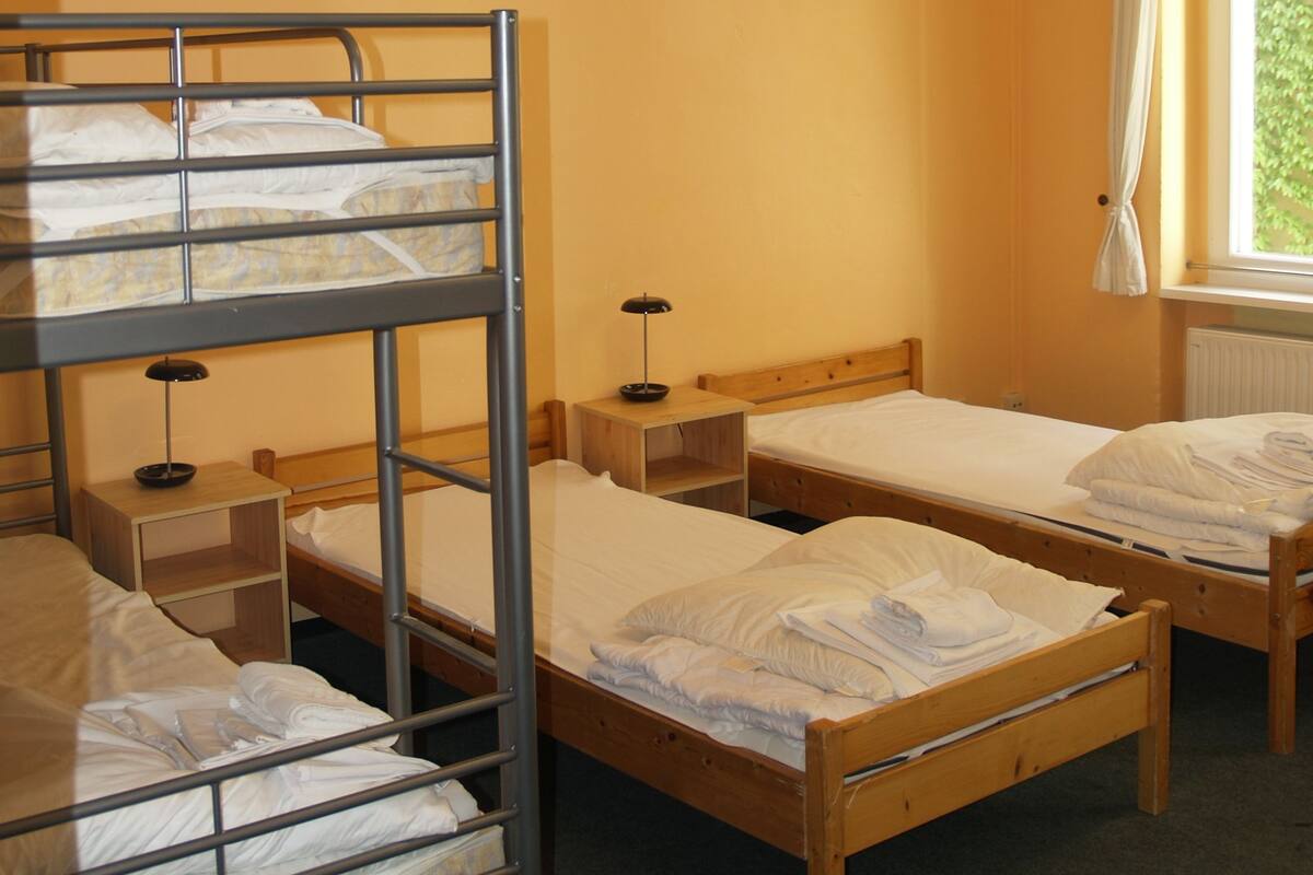 A dormitory room with bunk beds, a wooden nightstand, a lamp, and a painting on the wall.