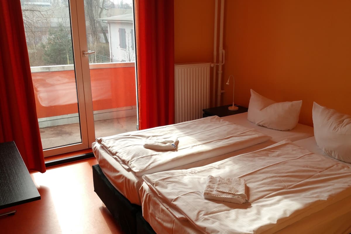 A double bed with white linens, a nightstand, and a view of a building through a window with red curtains.