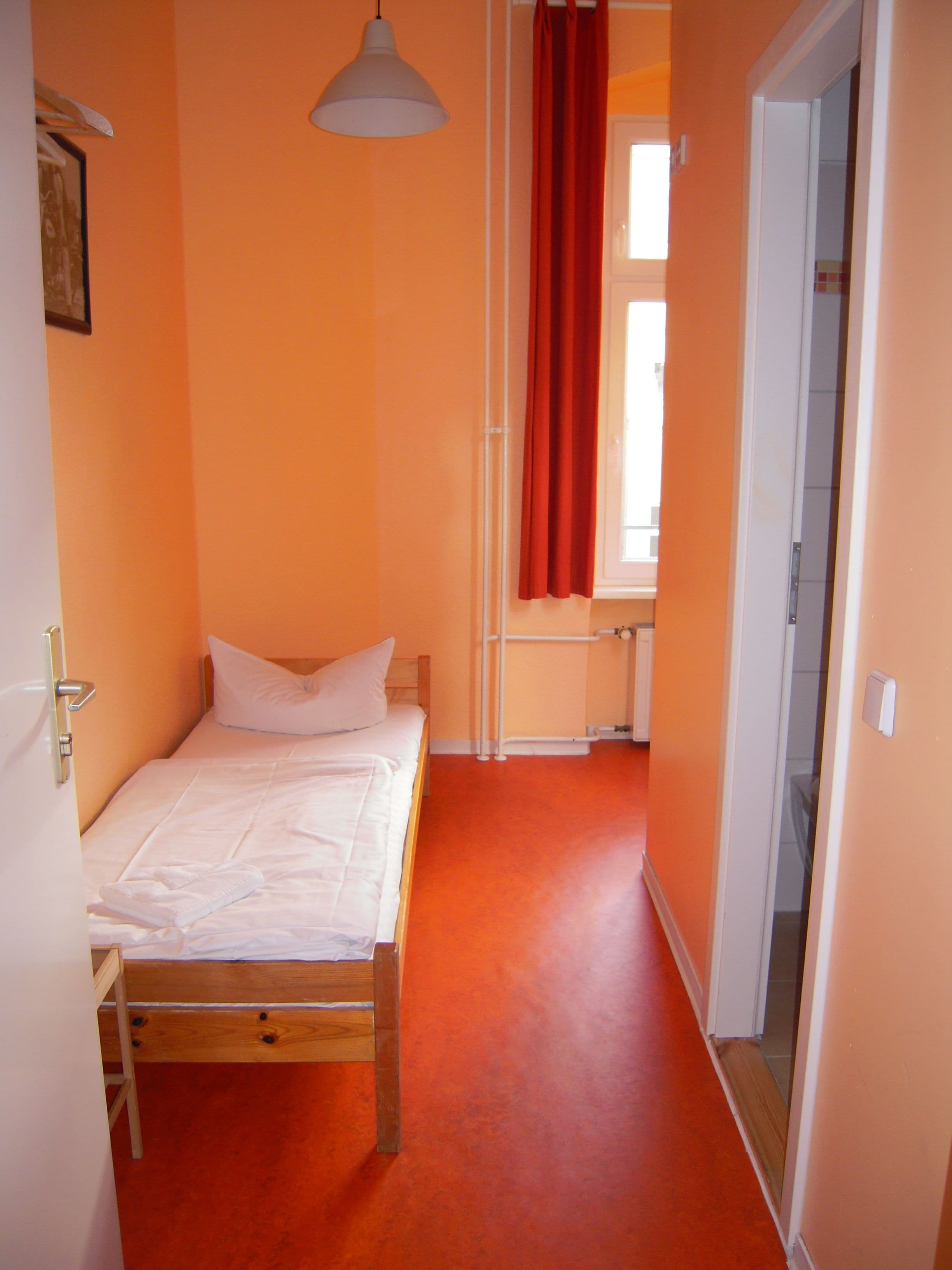 A small, single-bed room with a wooden bed frame, white bedding, and a red curtain.