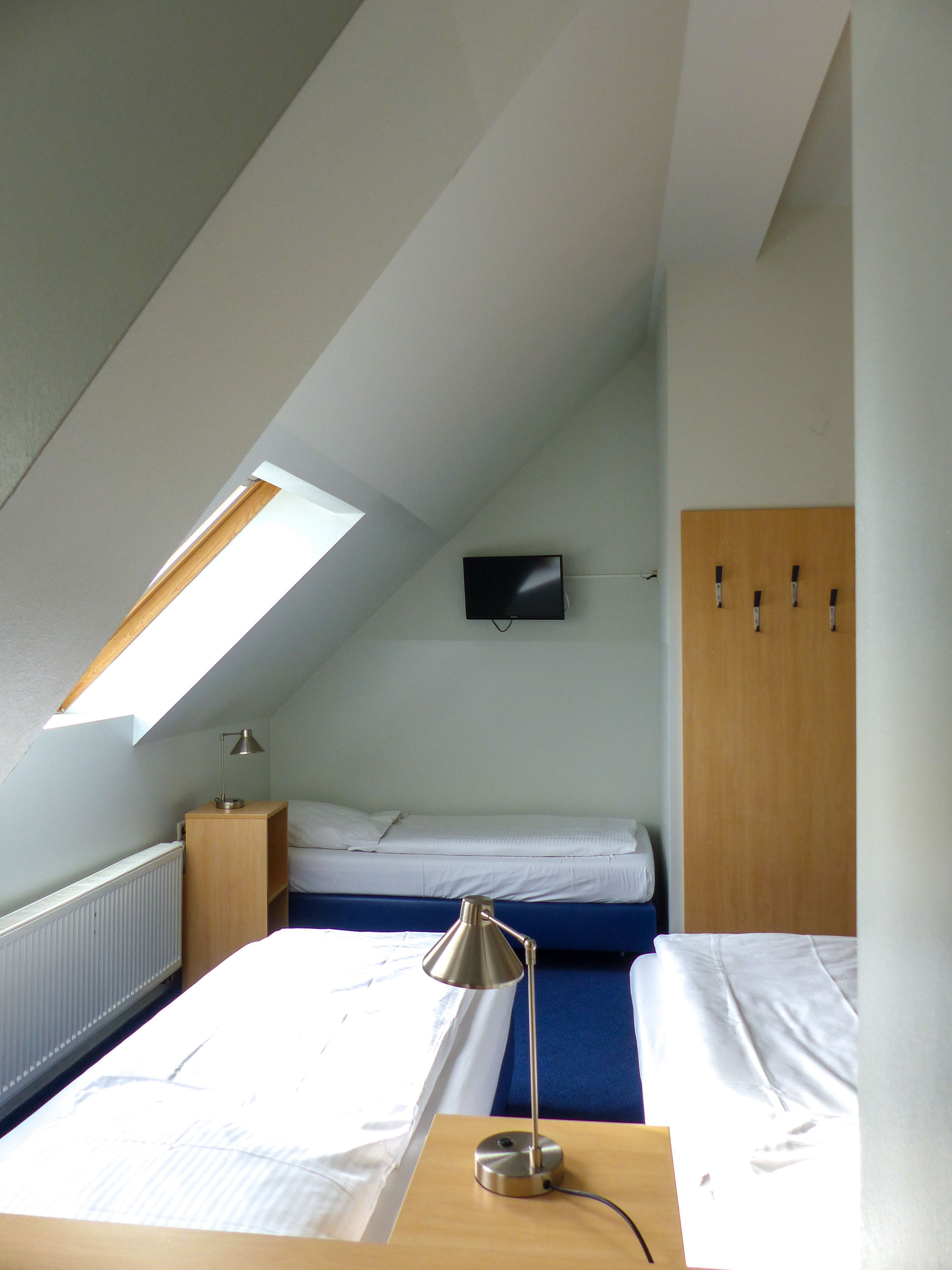 A dorm-style room with a bunk bed, a desk with a lamp, a TV mounted on the wall, and a wooden wardrobe.