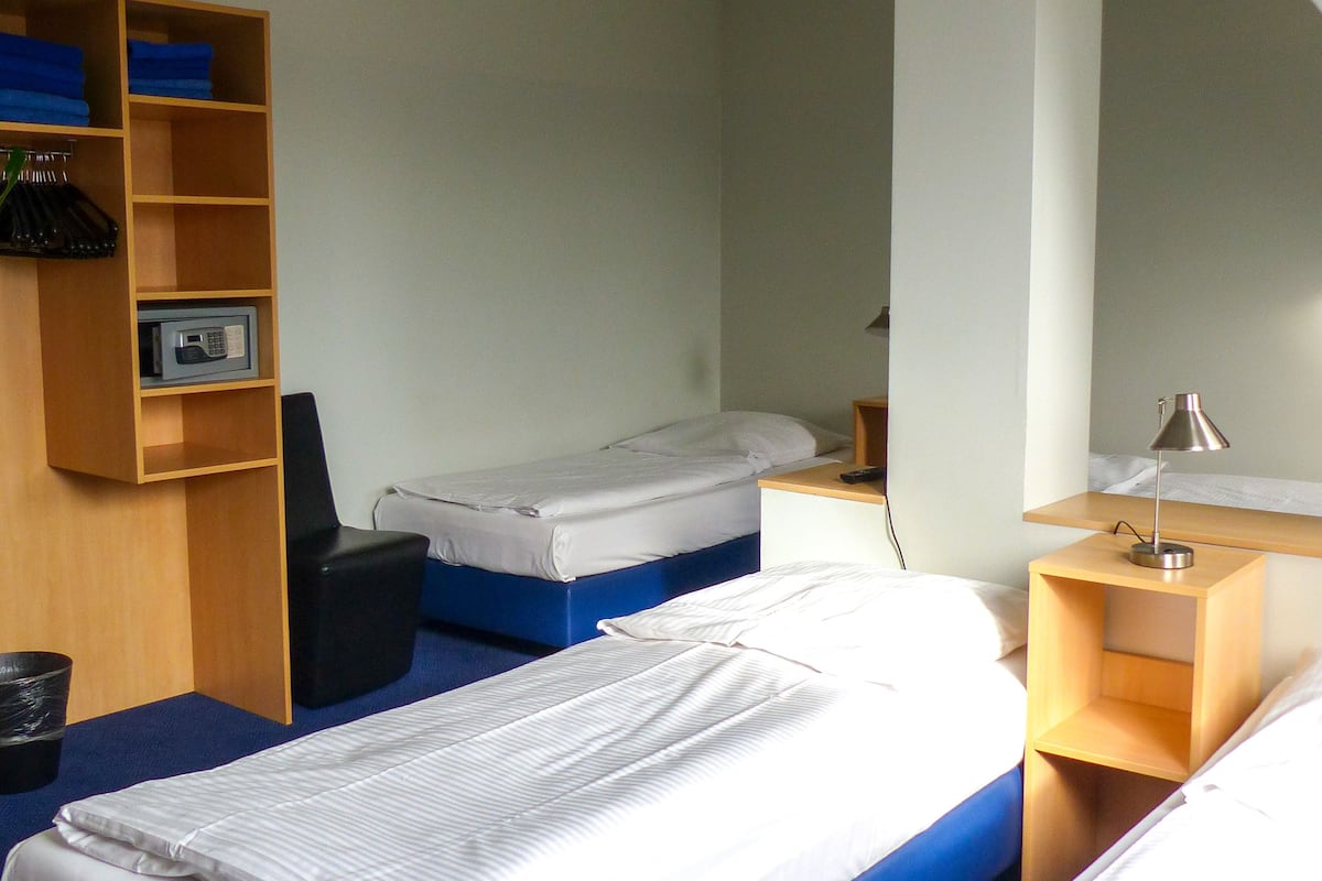 Quarto de hotel com duas camas, uma escrivaninha, uma cadeira, uma prateleira com roupas dobradas e uma mesinha de cabeceira com abajur.