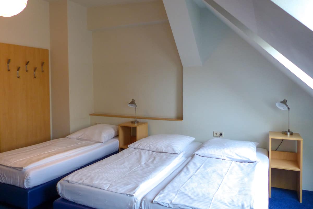 Quarto pequeno e moderno com duas camas de solteiro, um guarda-roupa de madeira e uma escrivaninha com luminária.
