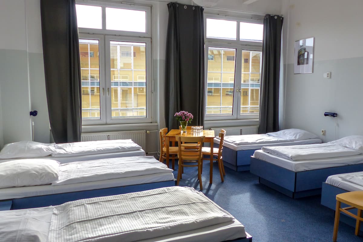 Quarto de hotel com quatro camas de solteiro, uma mesinha com um vaso de flores e janelas amplas com cortinas.