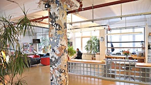 A spacious lobby with a high ceiling, exposed pipes, and a large mosaic column. There is a reception desk with a person behind it, and a variety of plants and seating areas. - Hotel Transit Loft Berlin (Berlin)