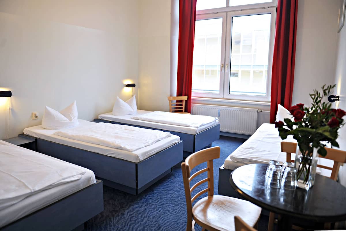 Quarto de hotel com duas camas de solteiro, uma mesinha com um vaso de flores e uma janela com cortinas vermelhas.