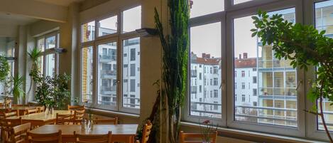 Un comedor con mesas y sillas de madera, ventanas amplias con vistas a edificios y plantas en macetas.