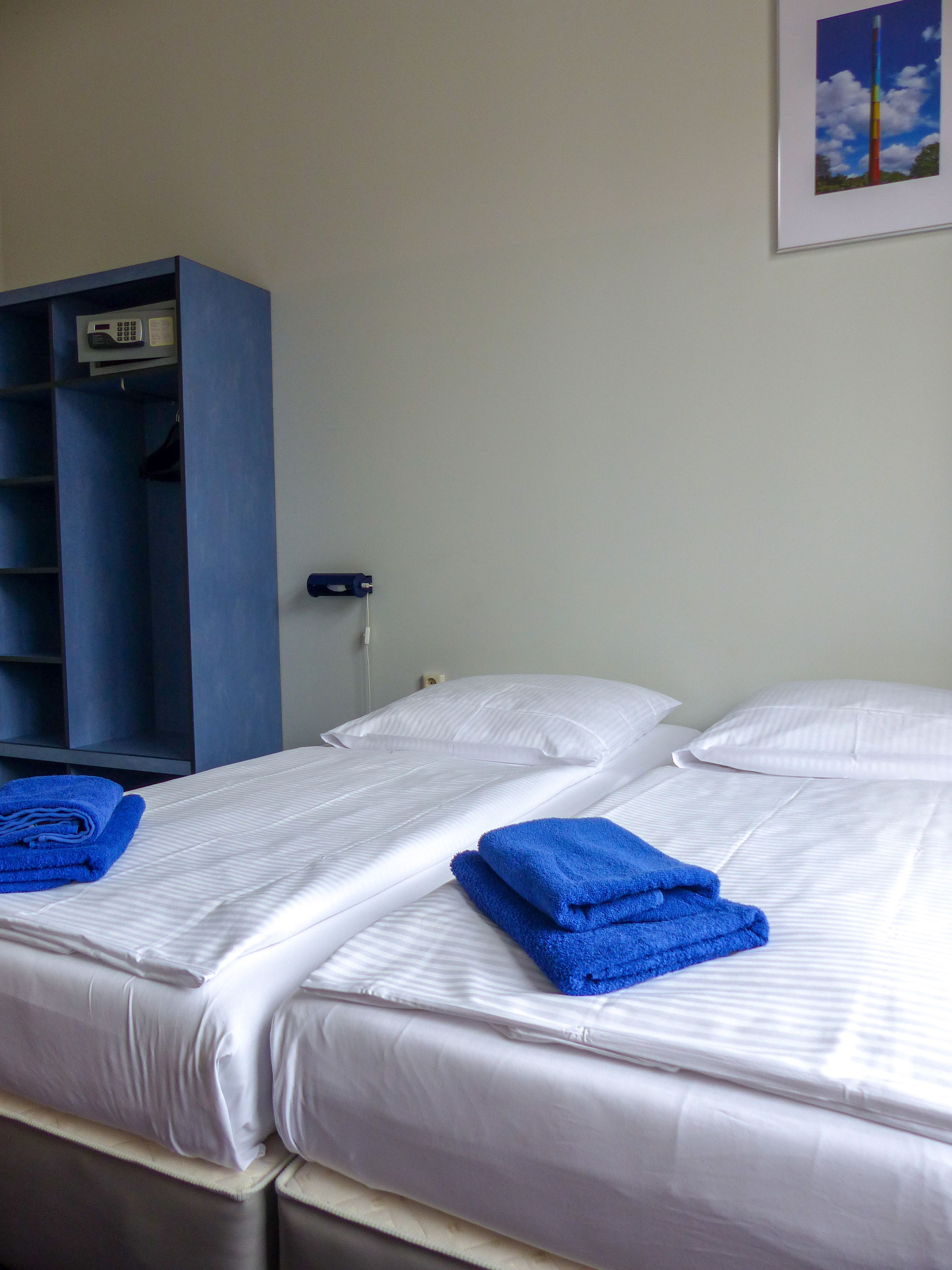 Une chambre d’hôtel avec deux lits, chacun étant équipé de draps blancs et de serviettes bleues. On y trouve également une armoire bleue et un téléphone fixé au mur.