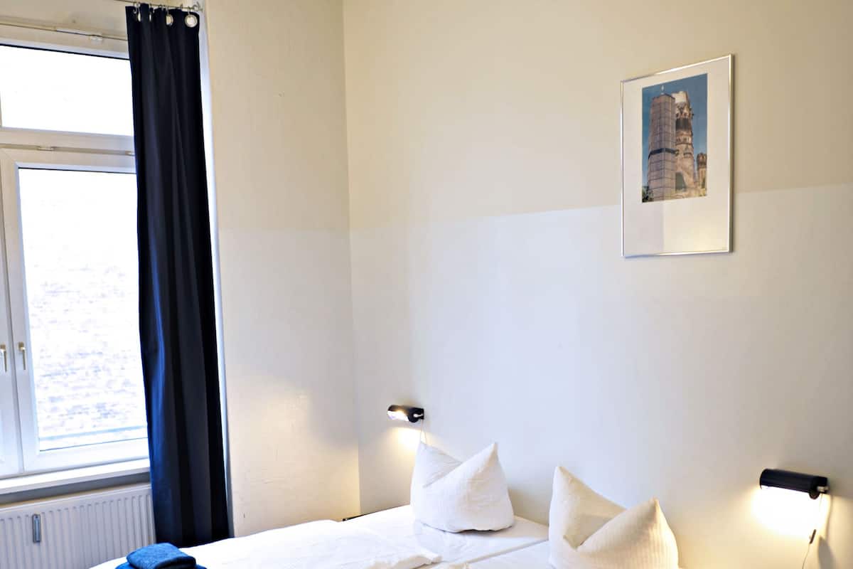 Quarto de hotel com duas camas, uma janela com cortinas, uma mesa de cabeceira com abajur e um quadro na parede.