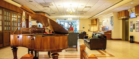 A grand piano in a spacious lobby with a chandelier, a reception desk, and a seating area.