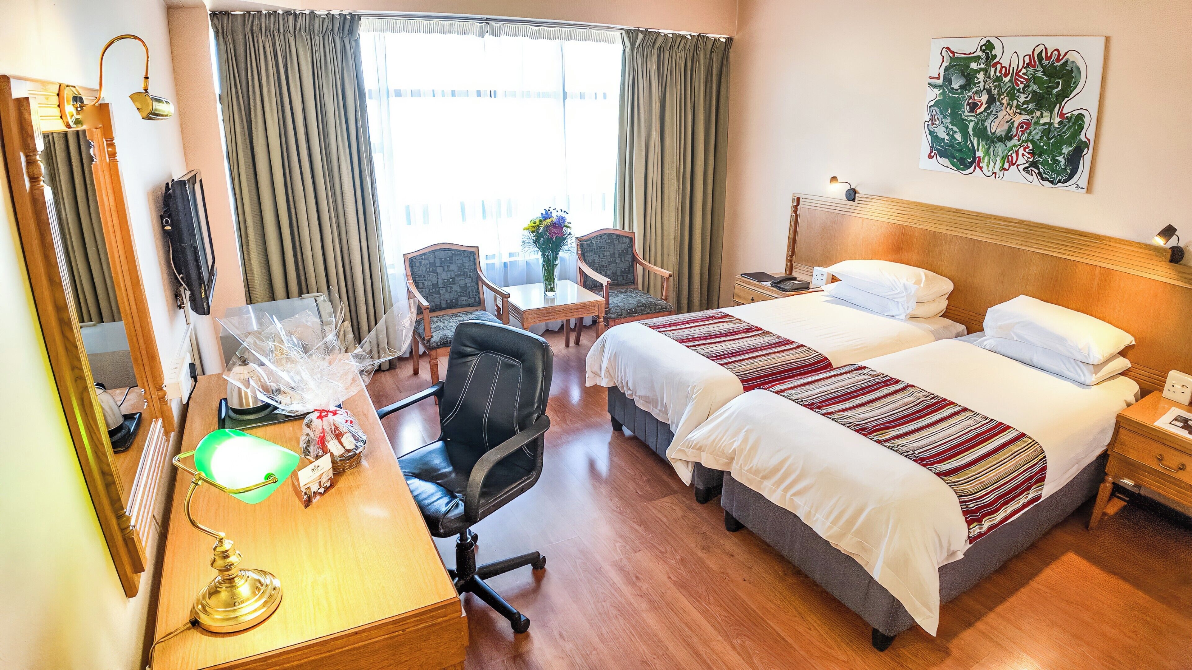 A hotel room with a desk, two beds, a chair, a table with a vase, and a painting on the wall.
